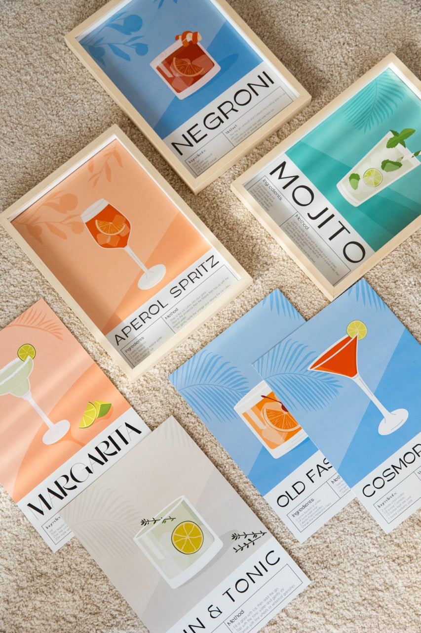 Yerde sergilenen, çerçeveli ve çerçevesiz Negroni, Mojito, Margarita, Aperol Spritz, Old Fashioned, Gin & Tonic ve Cosmopolitan içeren renkli kokteyl tarif posteri seti