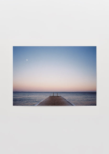 Gün batımında denize uzanan ahşap iskele, pastel gökyüzü ve ufukta görünen ayı betimleyen minimalist deniz fotoğrafı.