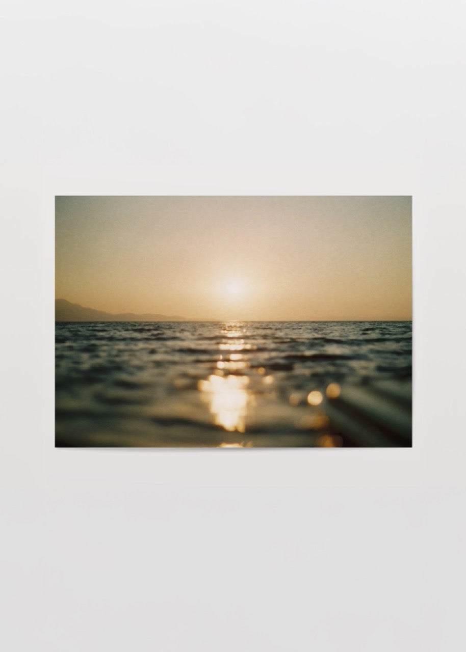 Deniz üzerinde gün batımının altın yansımalarını ve ufukta batan güneşi gösteren sıcak tonlu minimalist sahil fotoğrafı.