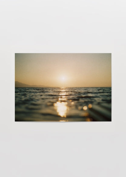 Deniz üzerinde gün batımının altın yansımalarını ve ufukta batan güneşi gösteren sıcak tonlu minimalist sahil fotoğrafı.