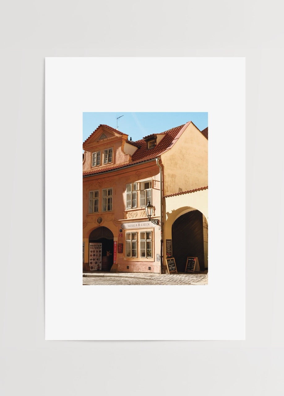 Merve Kurt imzalı Yaşayan Bina, Prag’da pastel pembe tarihi bina, kırmızı kiremit çatı, kemerli geçit ve klasik sokak lambasını gösteren mimari fotoğraf posteri.